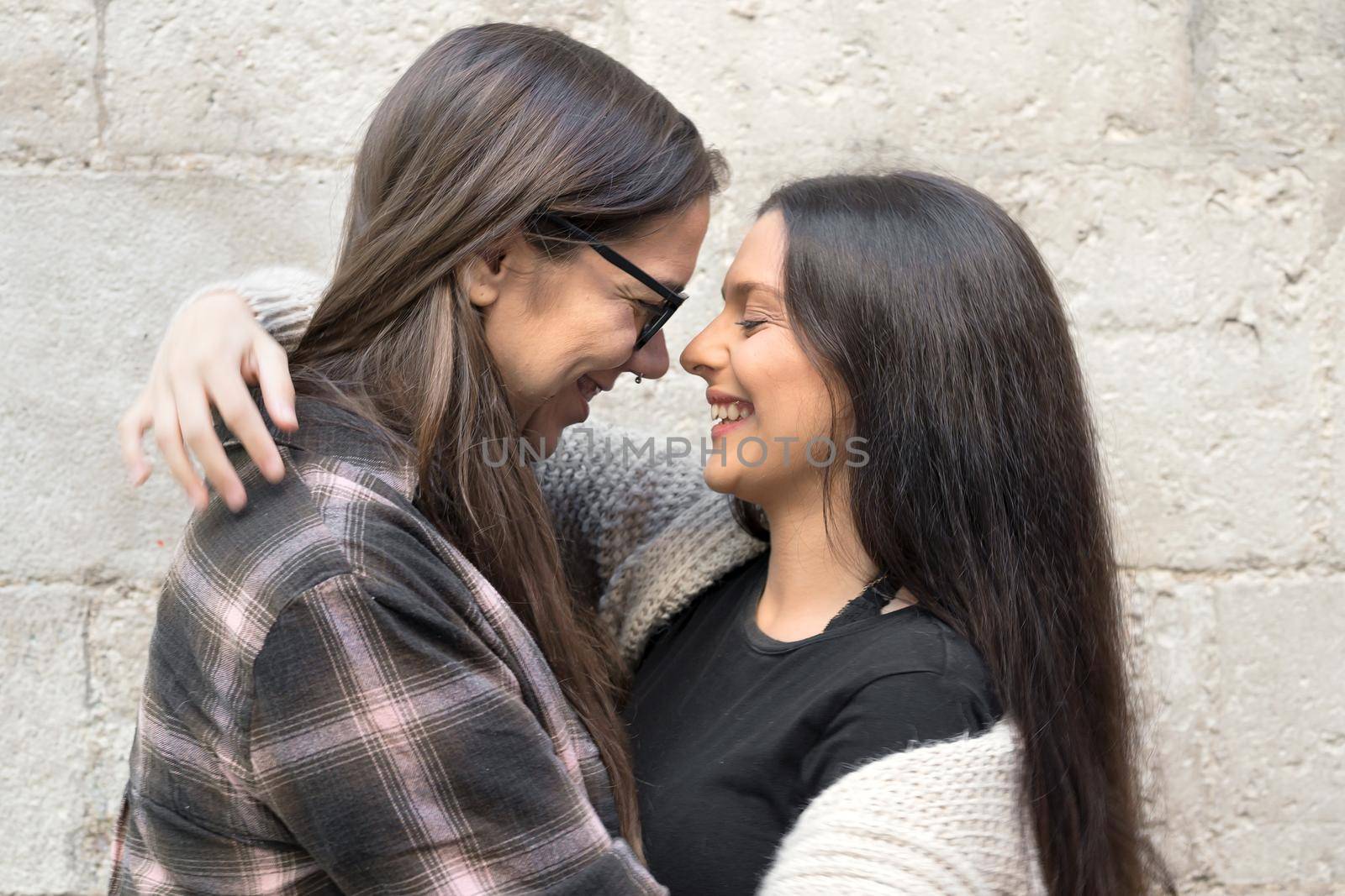 Portrait of an affectionate young lesbian couple hugging each other while standing together in front of a stone wall outside by HERRAEZ
