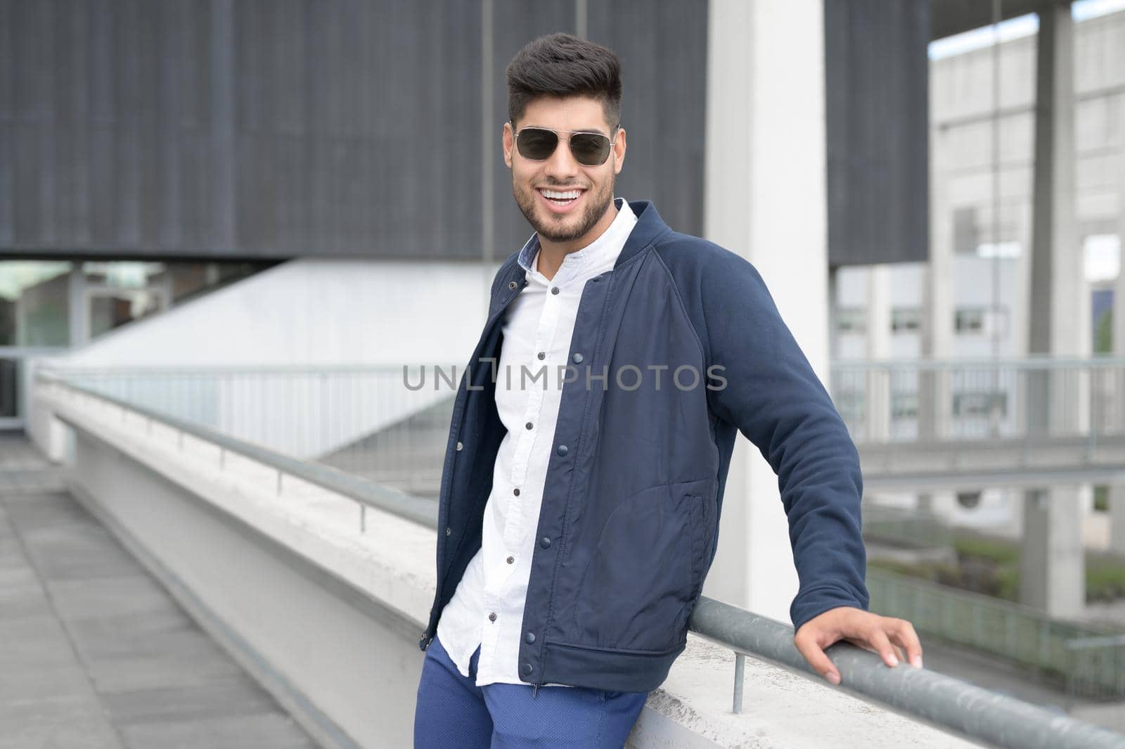 Young casual man looking at camera, wearing sunglasses is standing in modern city downtown. High quality photo.