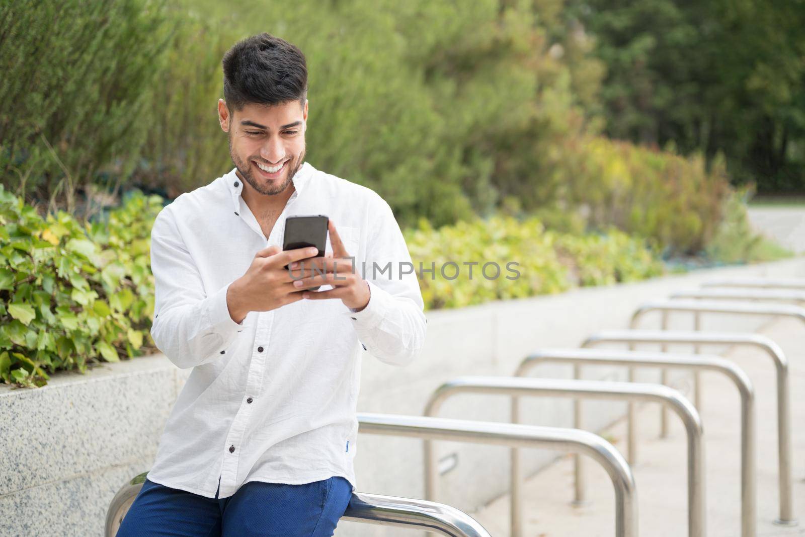 Handsome young man, smiling, using a mobile phone outdoors. by HERRAEZ