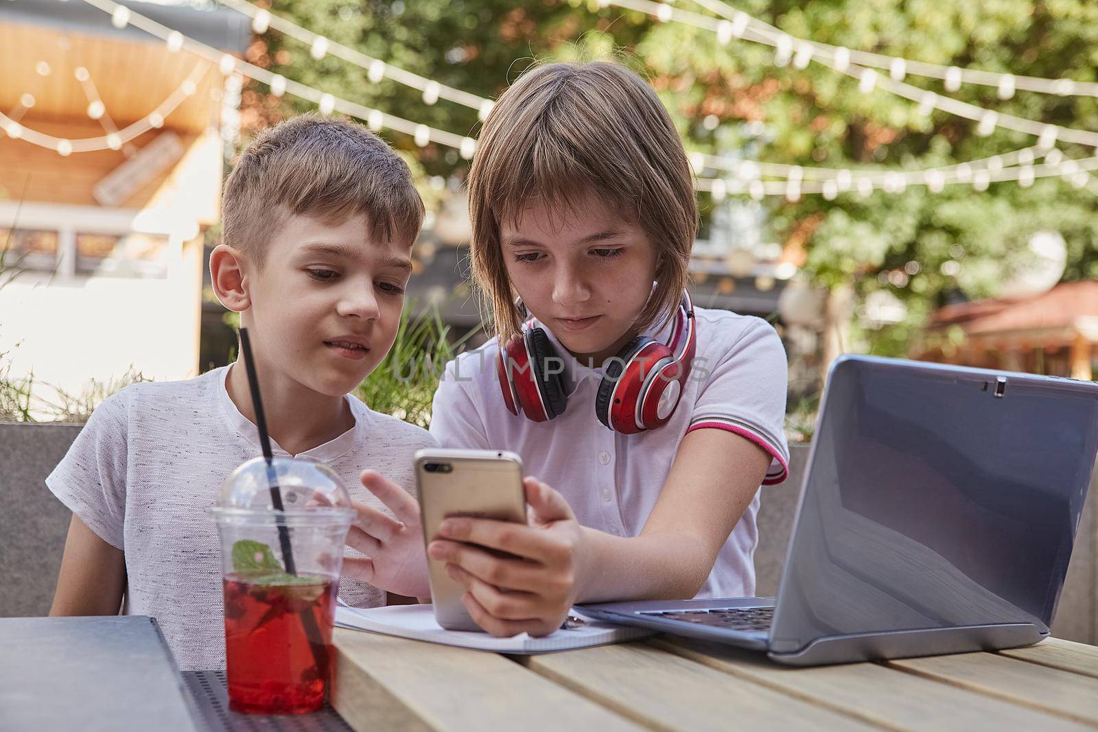 boy and girl sit outdoors wth laptop, watch at screen of smartphone. little caucasian schoolchildren sitting at table at cafe veranda, using mobile phone. modern technology, gadgets, digital devices