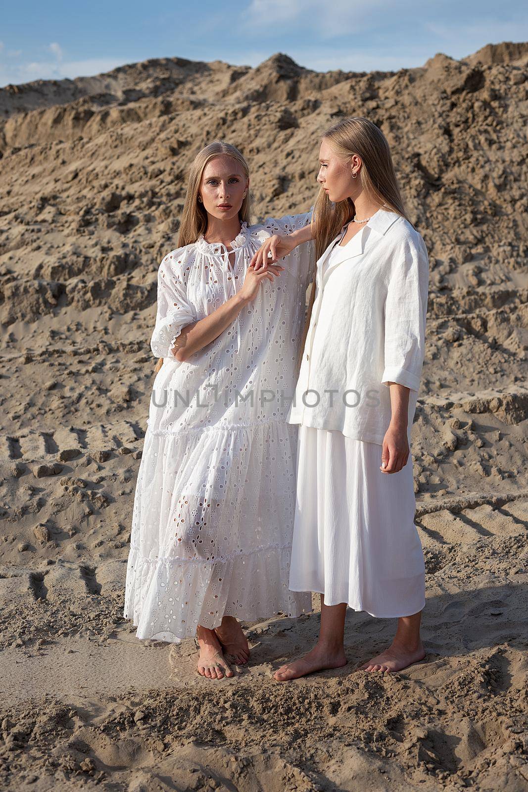 two attractive young twin sisters posing in sand quarry in elegant white clothes by artemzatsepilin