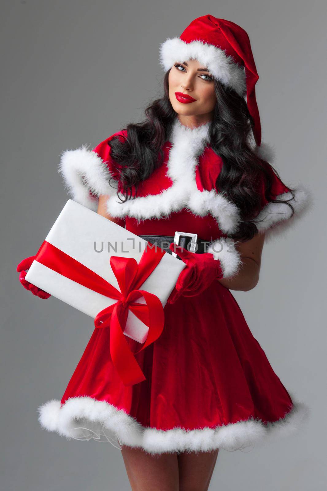 Beautiful young woman Santa Claus helper in red dress and hat holding gift box, white background, merry christmas happy holidays