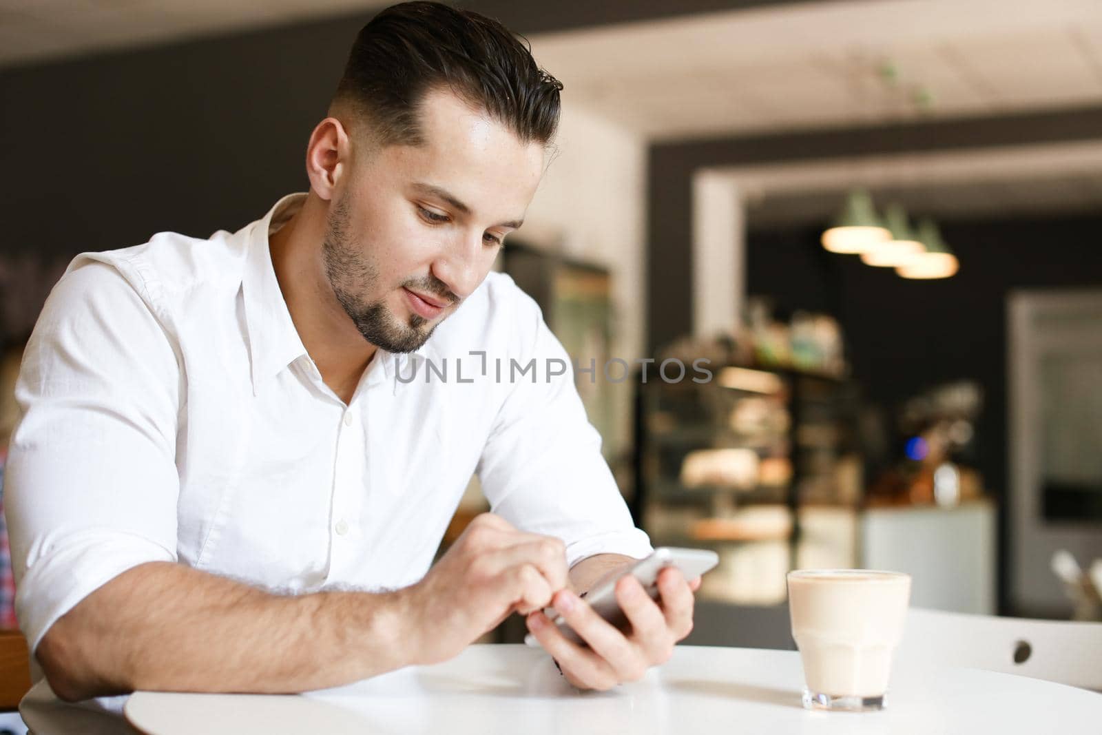 Young businessman chatting by smartphone at cafe, wearing white shirt. Concept of business and modern technology, free hotspot.