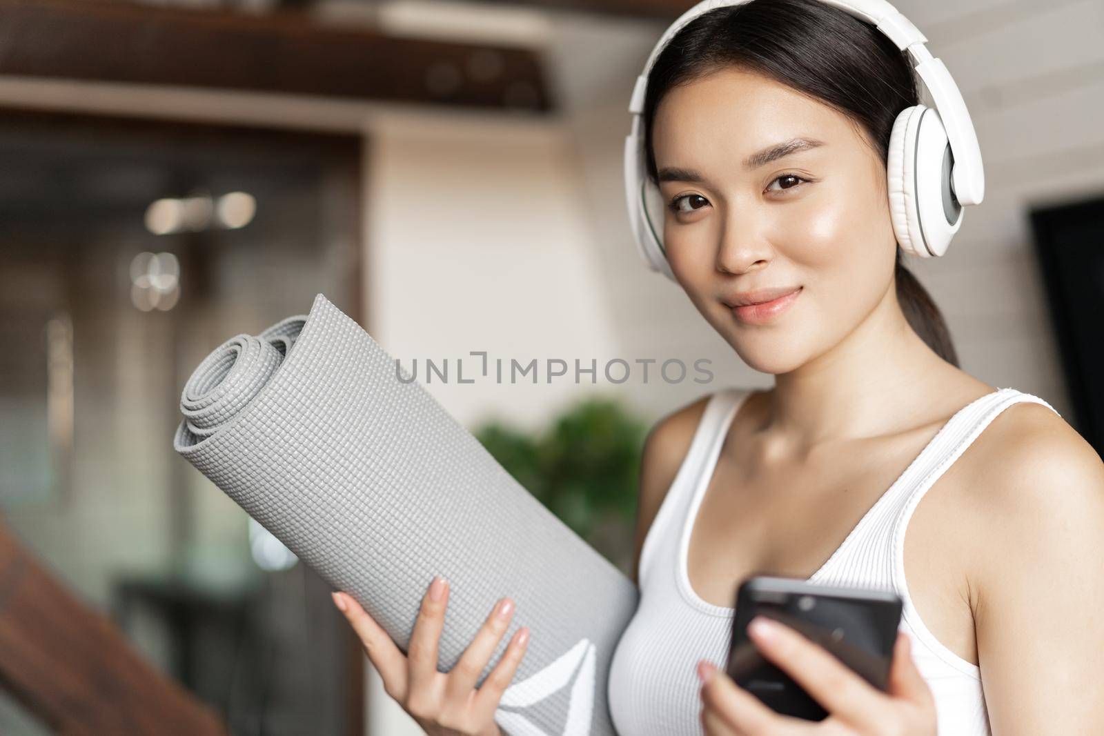 Smiling asian girl in headphones, listening music at home and workout with floor mat, holding mobile phone, fitness exercises in living room.