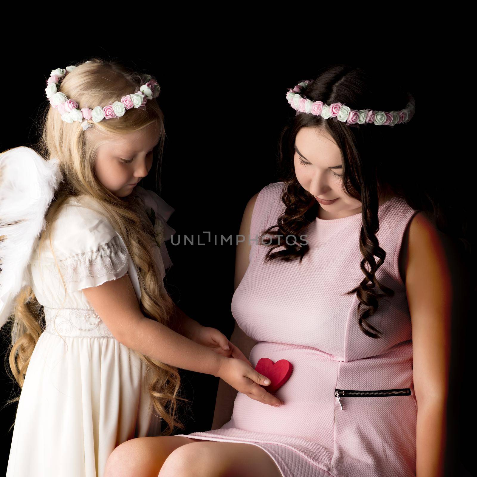 A little girl in the image of an angel hugs the belly of her eldest pregnant sister. The concept of family happiness, the birth of a child.