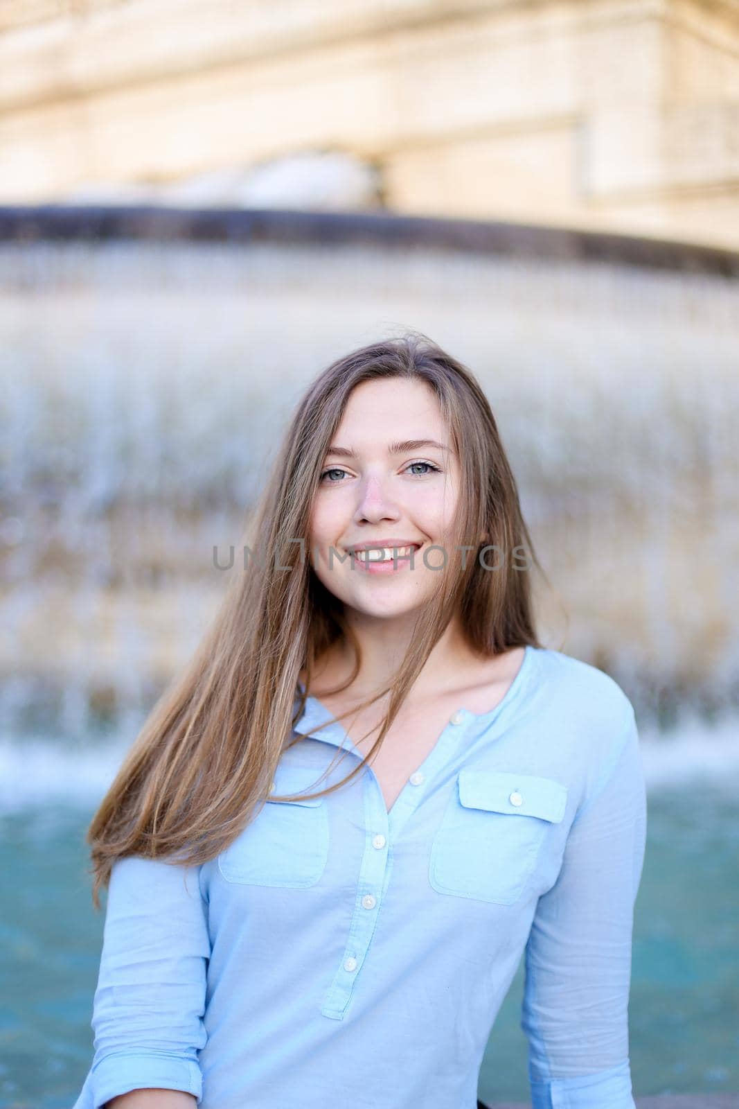 Portrait of young caucasian student sitting near fountain and smiling, wearing blue shirt. Concept of tourism and urban resting.