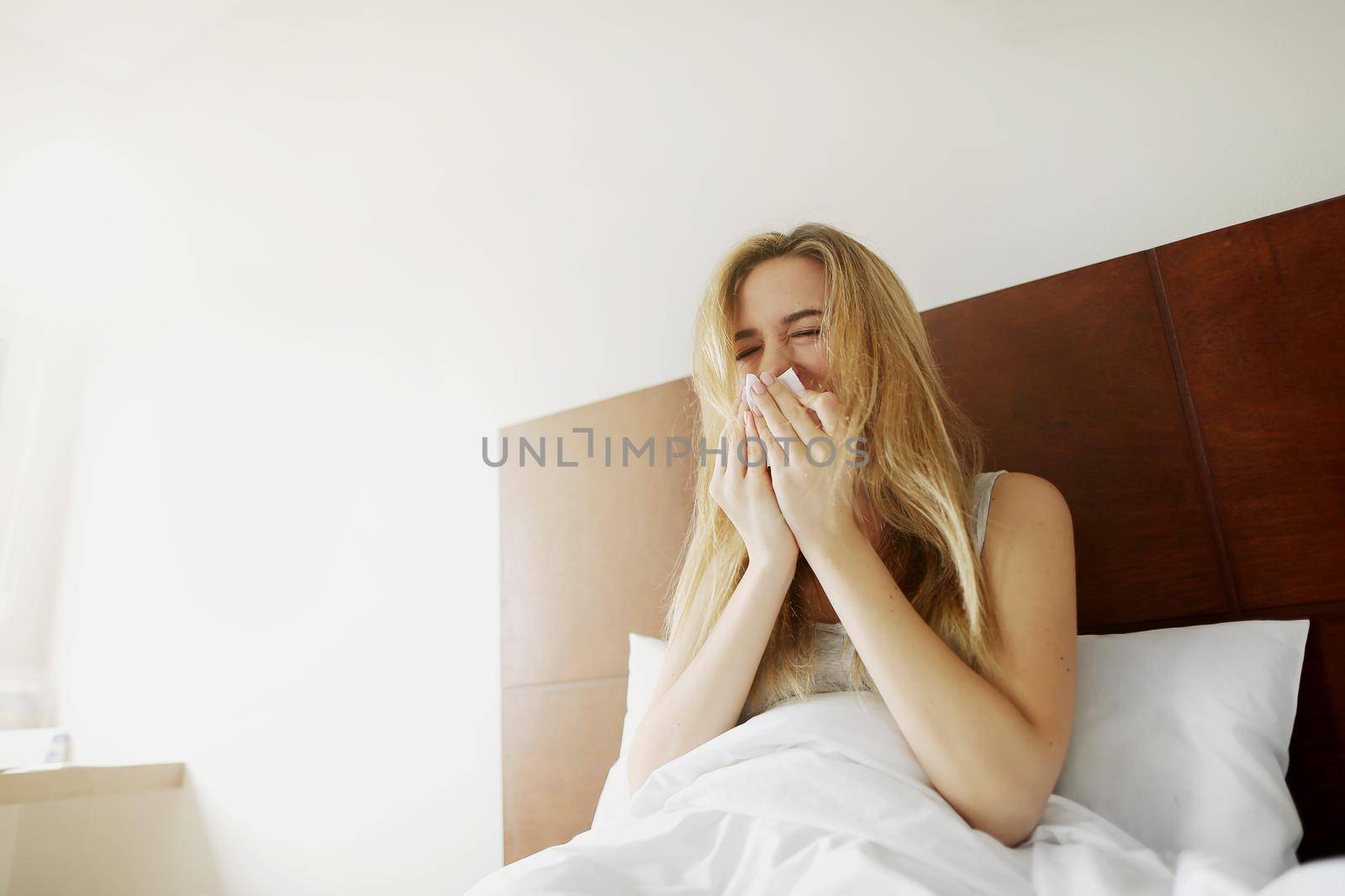 ill Sunlight Female american woman sneezes with napkin in white bed , get cold outside. Concept of pills medical treatment advertisement