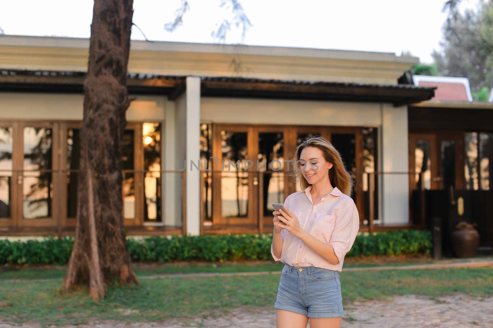 Young european female peson typing message by smarptone near resort house, wearing blouse and shorts. Concept of modern technology and summer vacations.