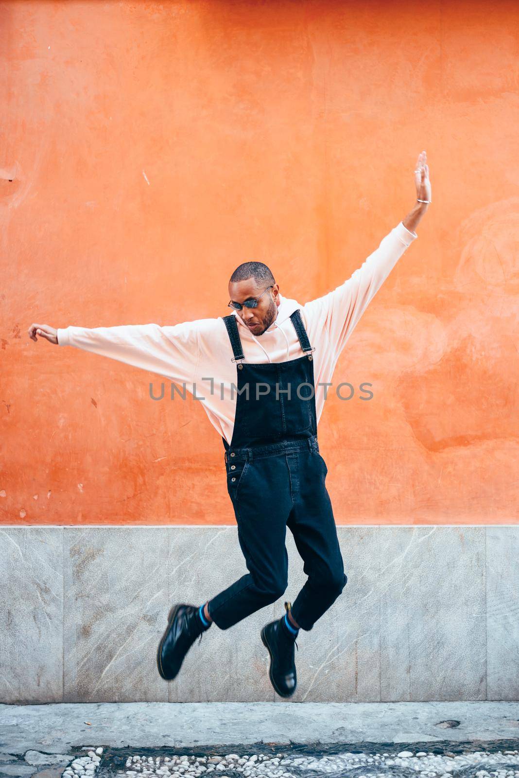 Young black man wearing casual clothes jumping in urban background. Lifestyle concept. Millennial african guy with bib pants outdoors