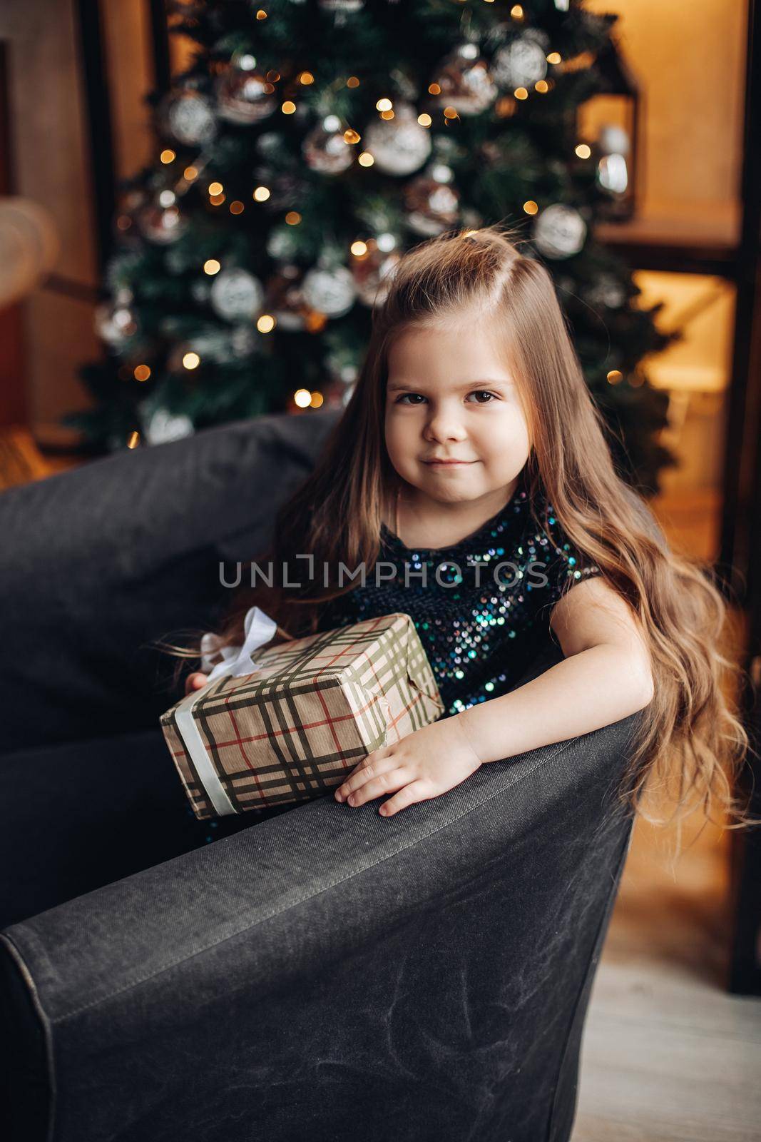 Stock photo portrait of a pretty little girl with long wavy fair hair wearing sparkling top, sitting in cozy arm-chair with wrapped Christmas present.