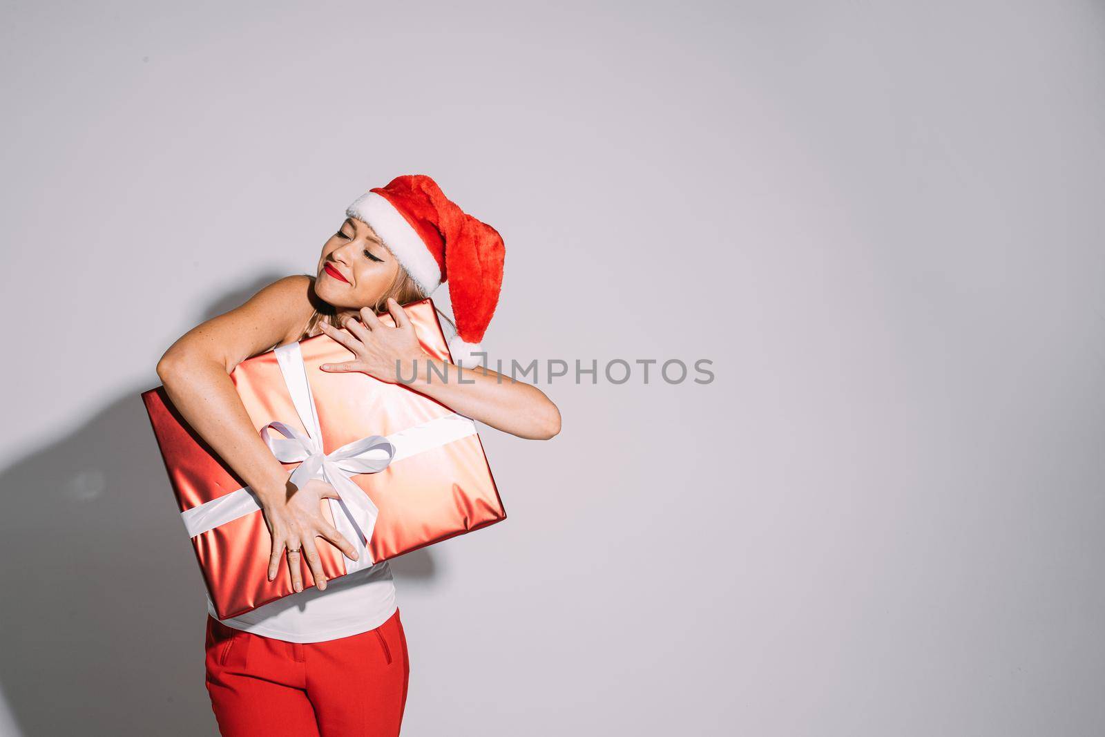 Happy young woman in Santa hat having her eyes closed while smiling and hugging a Christmas present. Holiday concept