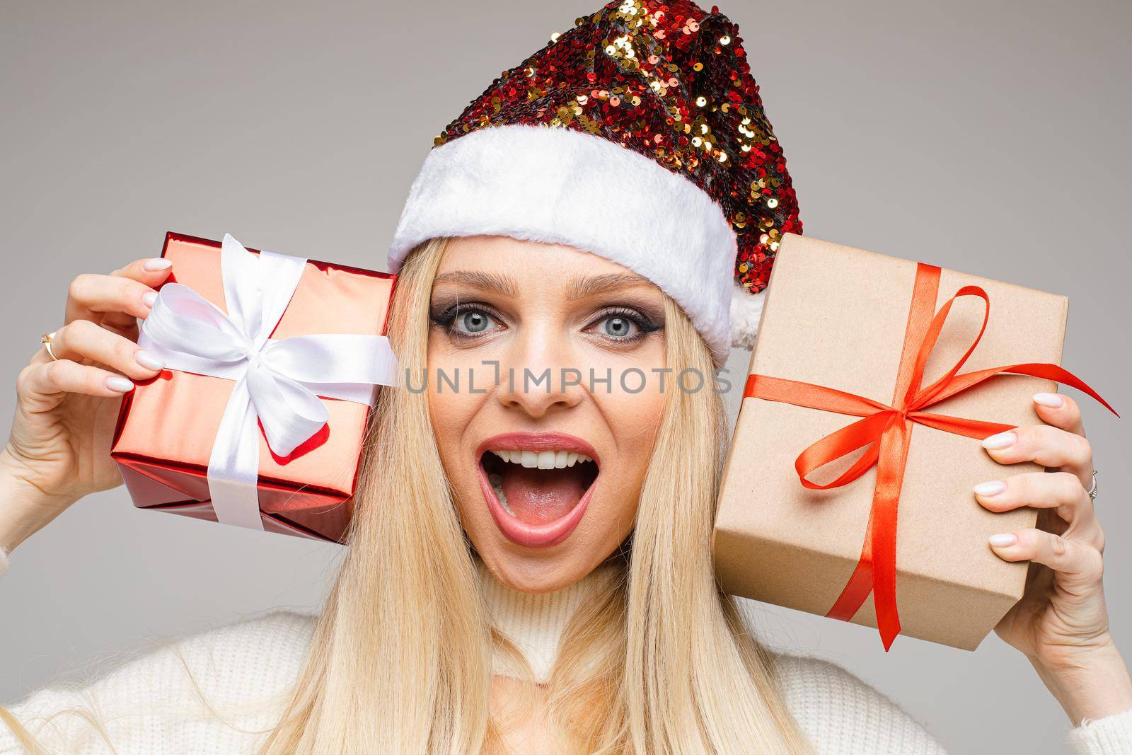 Close up of smiling pretty lady wearing Santa hats and posing with presents, isolated on grey background. New Year holiday concept