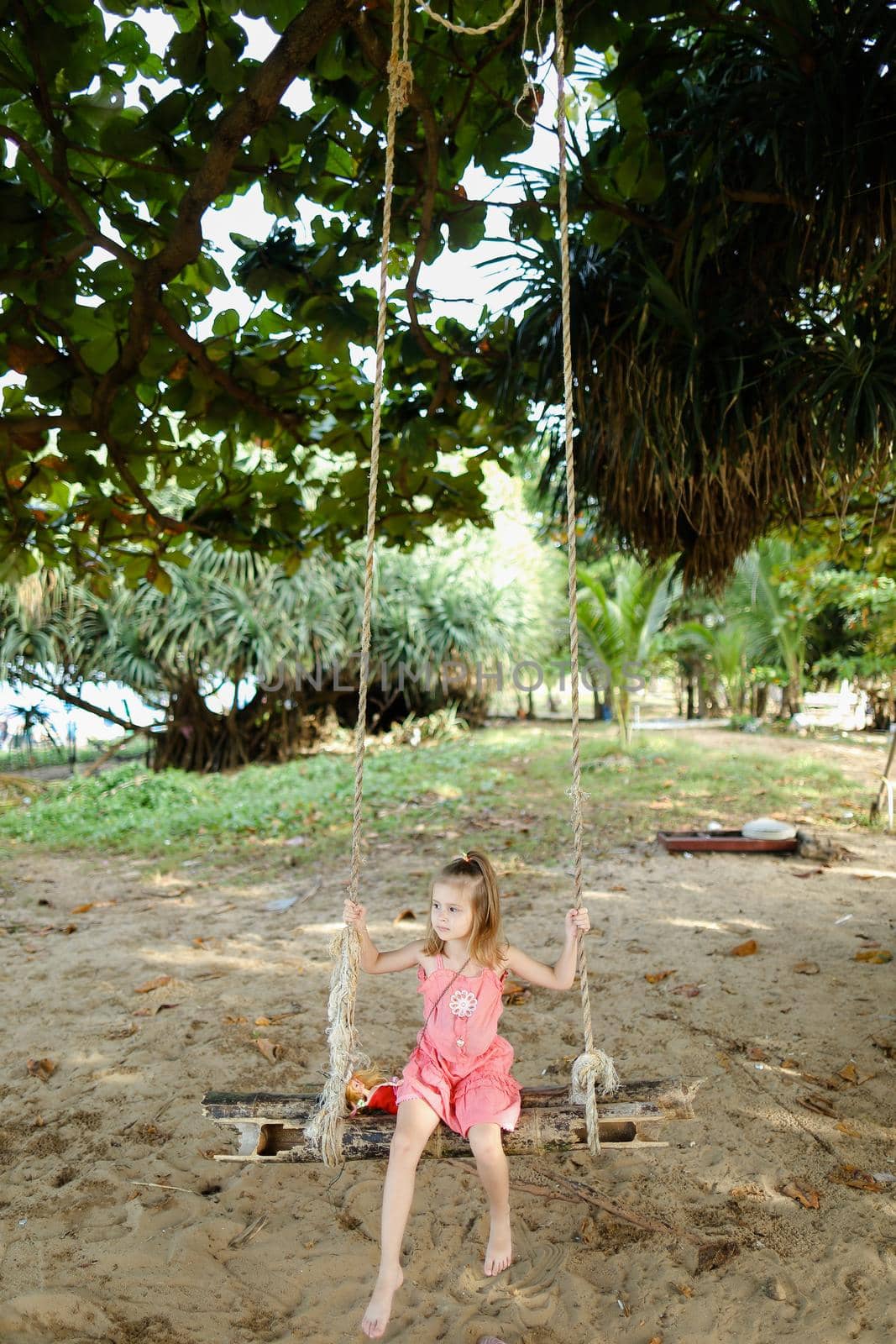 Little funny girl riding on swing on sand background, wearing pink dress. Concept of childhood and summer vacations on exotic resort.