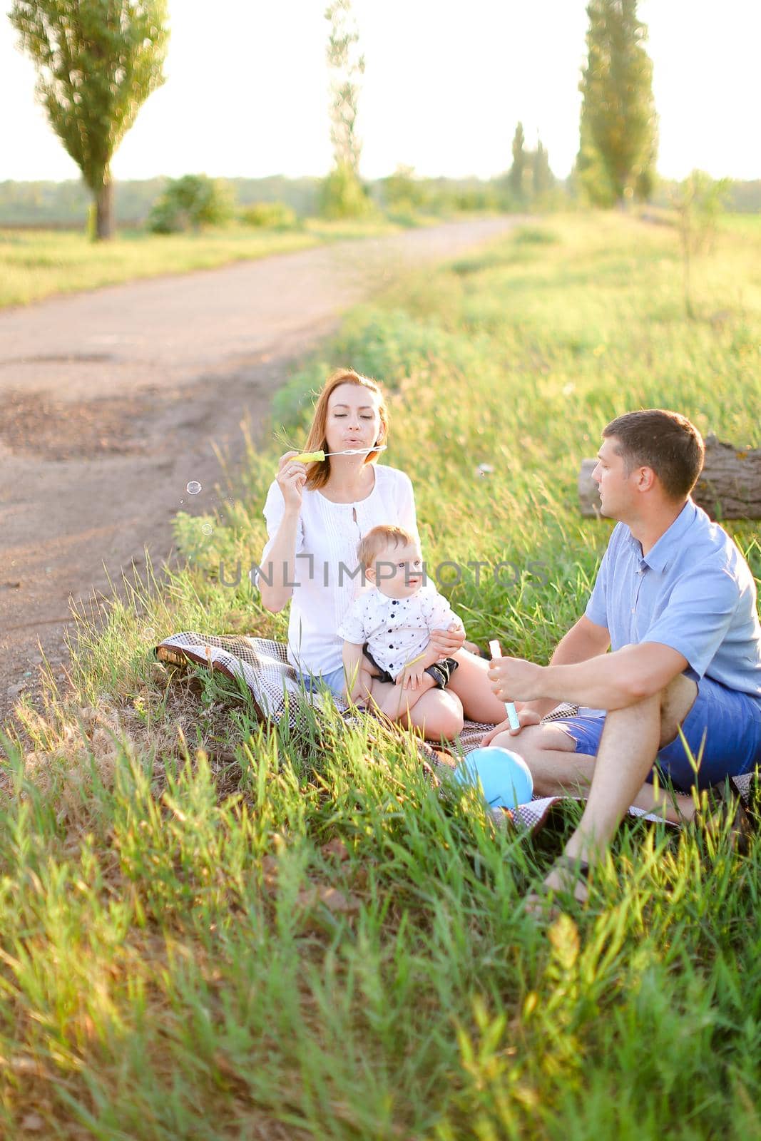Young Caucasian father and blonde mother sitting on grass with little baby and blowing bubbles. Concept of picnic with child and resting on nature.