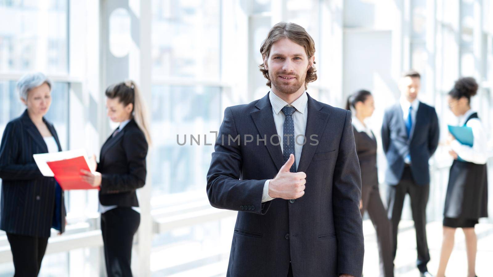 Hansome young businessman in formal suit standing with thumb up in front of colleagues in business building