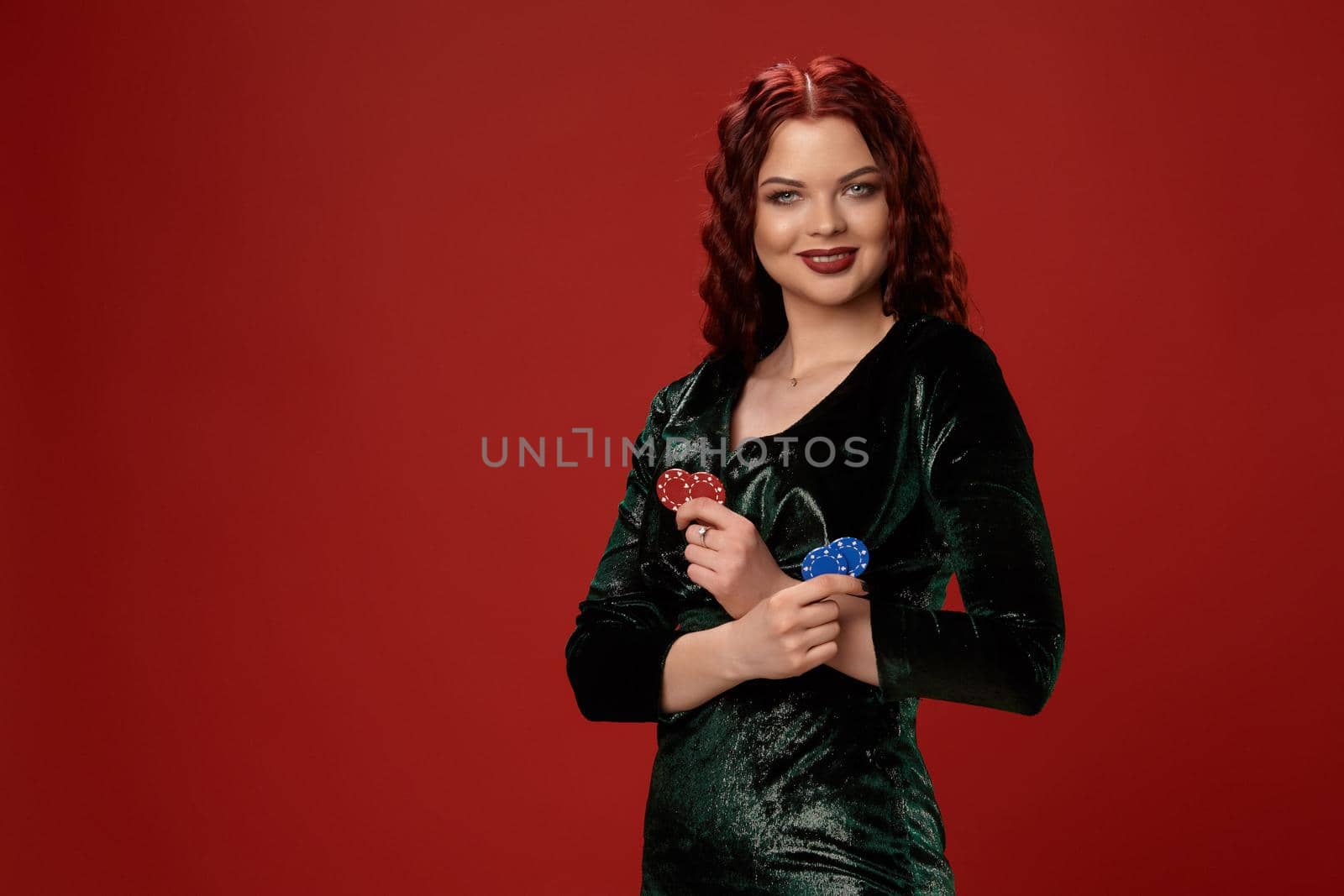 Awesome young redheaded woman is holding some gambling chips in her crossed hands and smiling, on a red background.