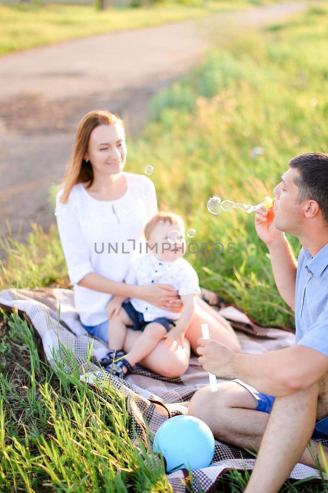 Young european mother and father sittling on grass with little baby and blowing bubbles. Concept of picnic and children, parenthood and leisure time.