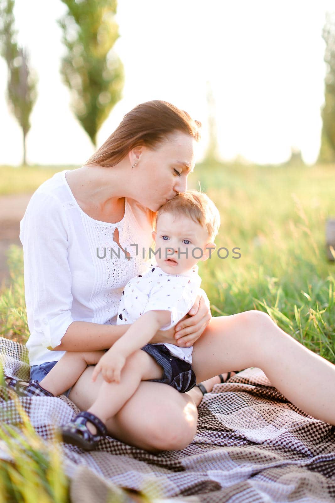 Young blonde woman sitting with little baby on plaid, grass on background. Concept of picnic, motherhood and nature.