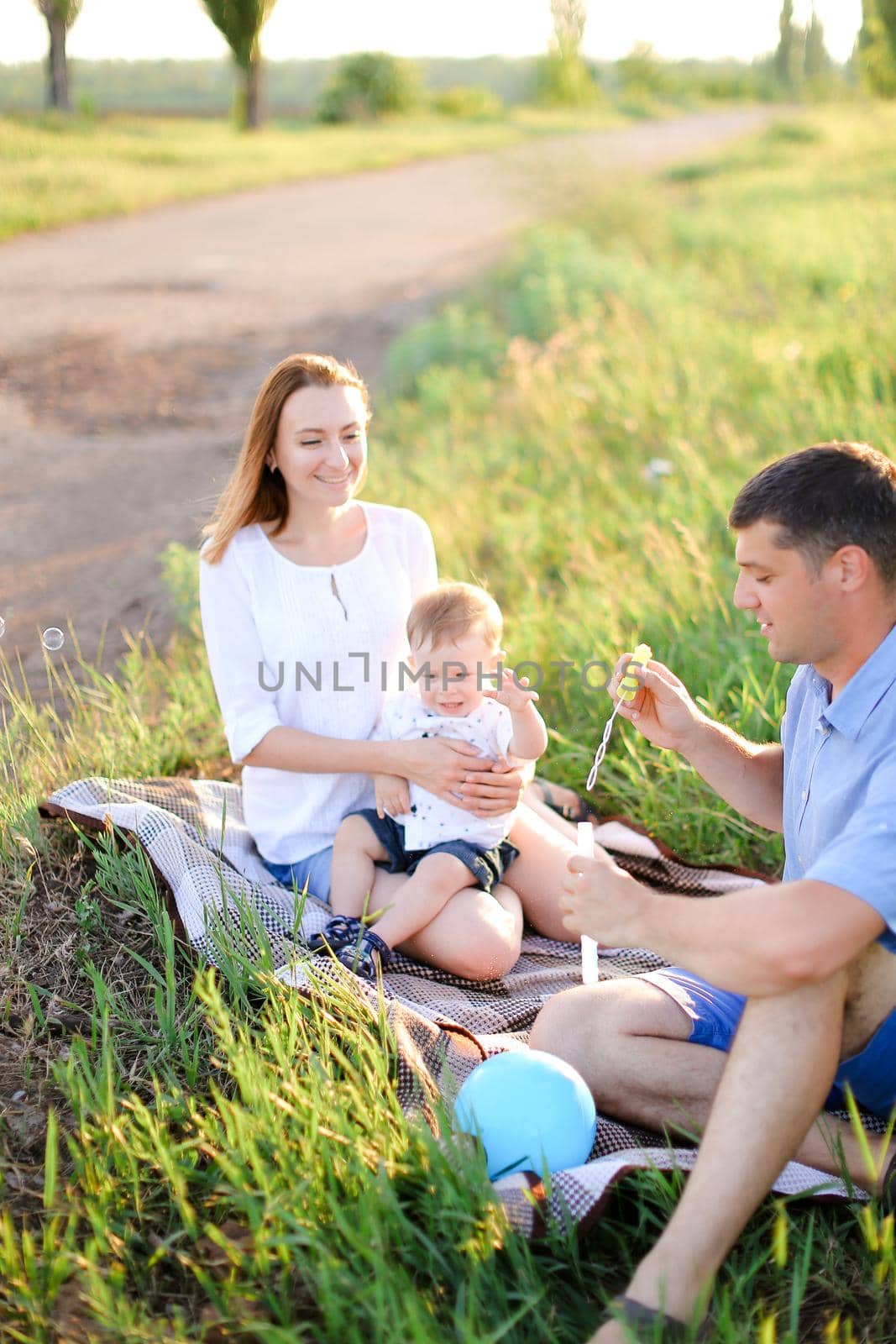 Young smiling mother and father sittling on grass with little baby and blowing bubbles. Concept of picnic and children, parenthood and leisure time.
