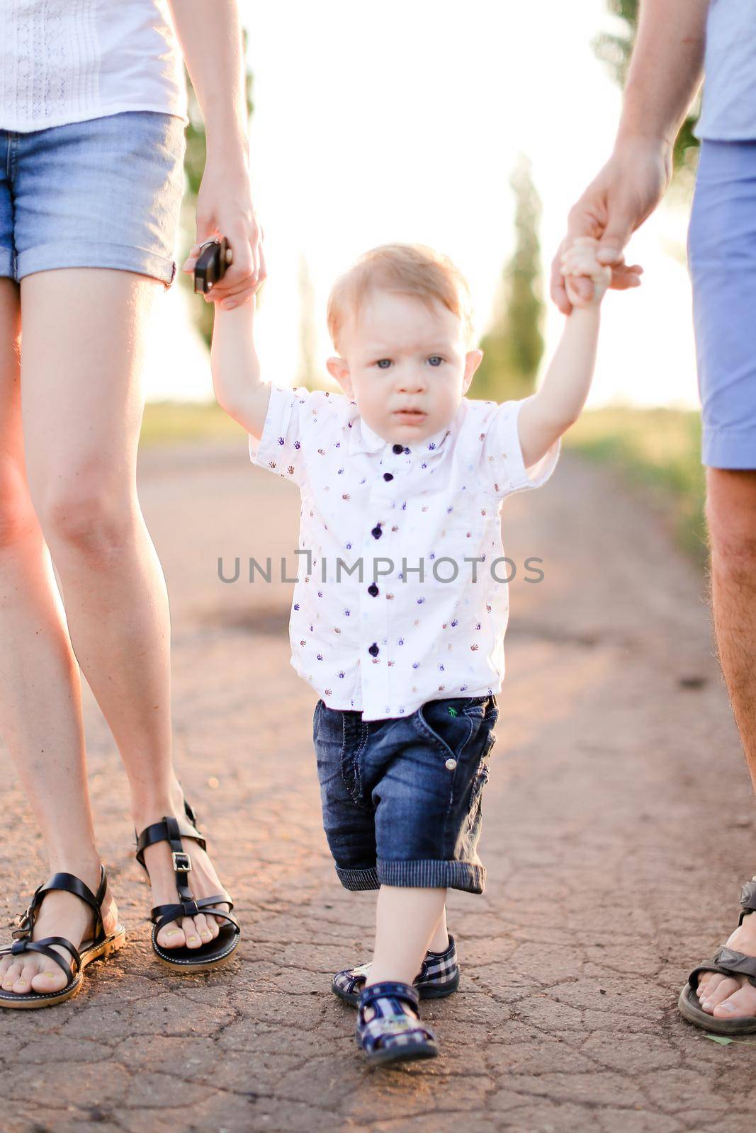 Little male child holding parents hands and walking on road. Concept of childhood and resting on open air.