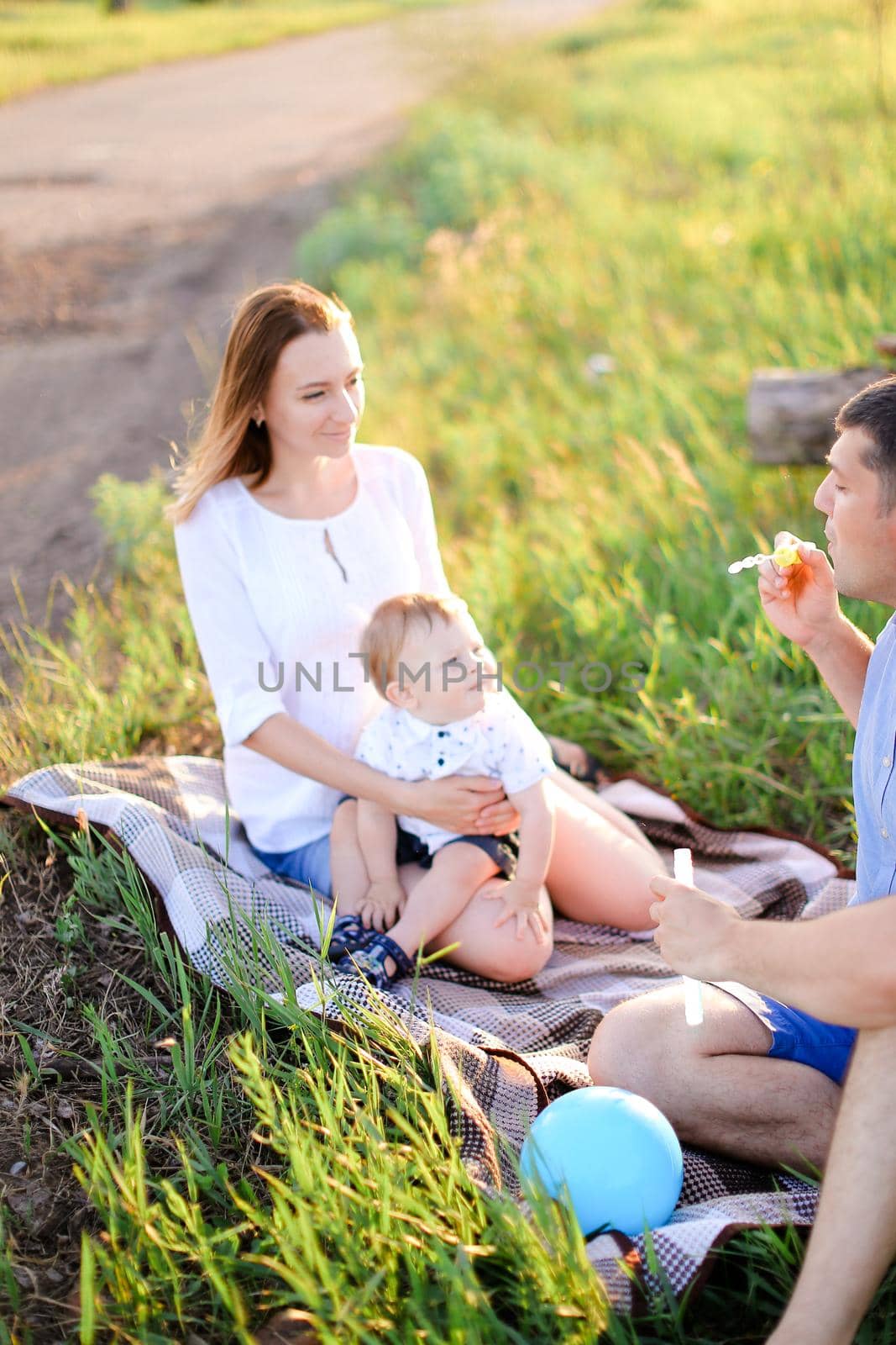 Young mother and father sittling on grass with little baby and blowing bubbles. Concept of picnic and children, parenthood and leisure time.