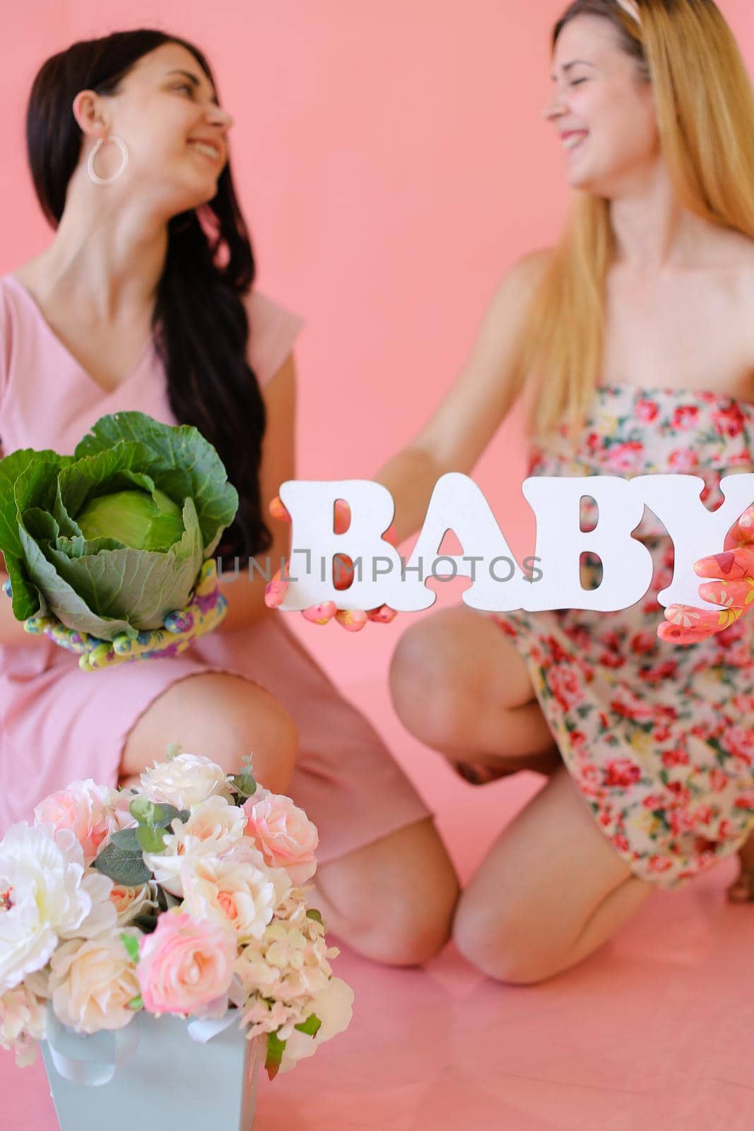 Young female persons keeping cabbage and inscription baby in pink monophonic backgrpund. Concept of photo session for pregnant mom.