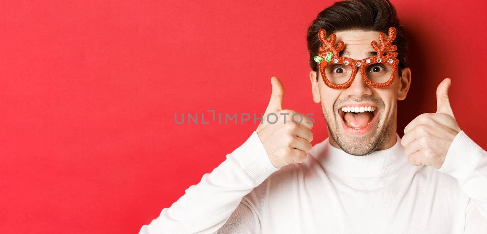 Concept of winter holidays, christmas and celebration. Close-up of excited handsome man in party glasses, smiling and showing thumbs-up in approval, standing over red background by Benzoix