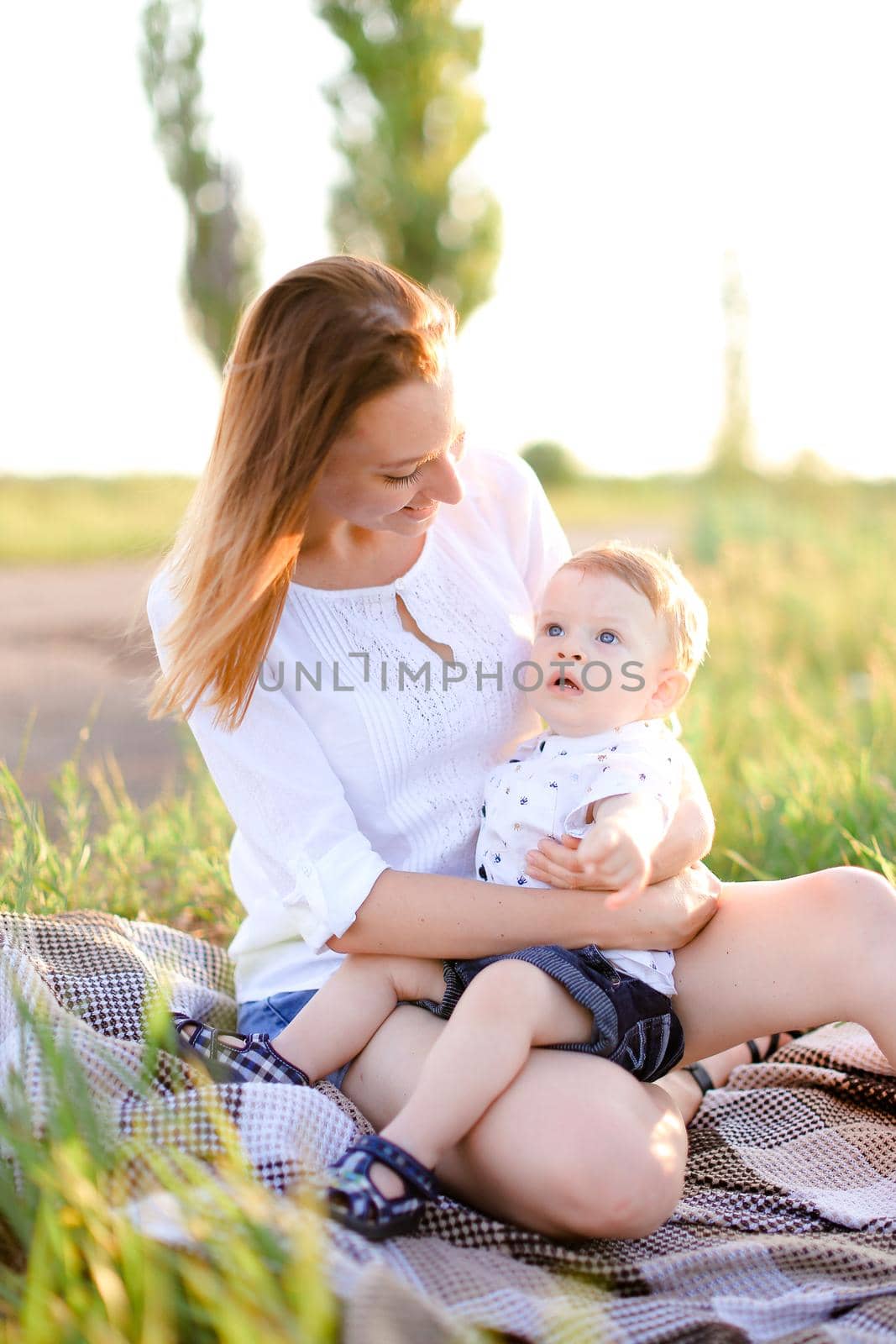 Young caucasian woman sitting with little baby on plaid, grass on background. Concept of picnic, motherhood and nature.