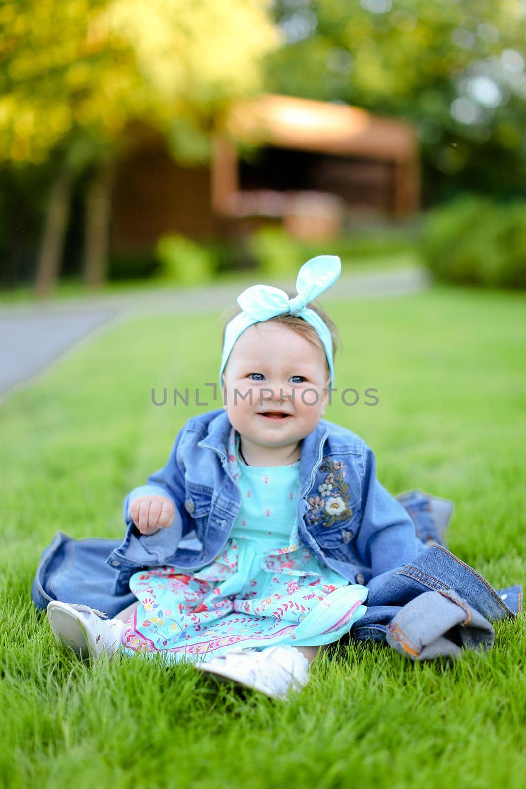 Little smiling female baby sitting on green grass and wearing jeans jacket. Concept of childhood and resting in garden, nature background.