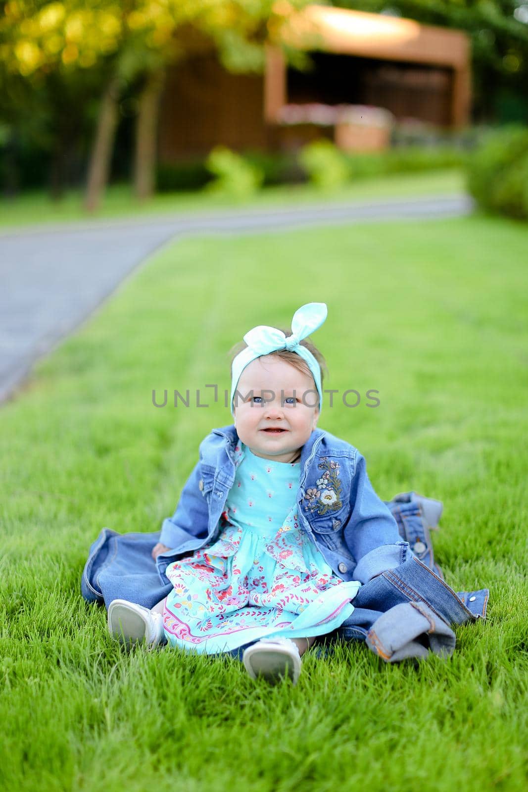 Little female baby sitting on green grass and wearing jeans jacket. Concept of childhood and resting in garden, nature background.