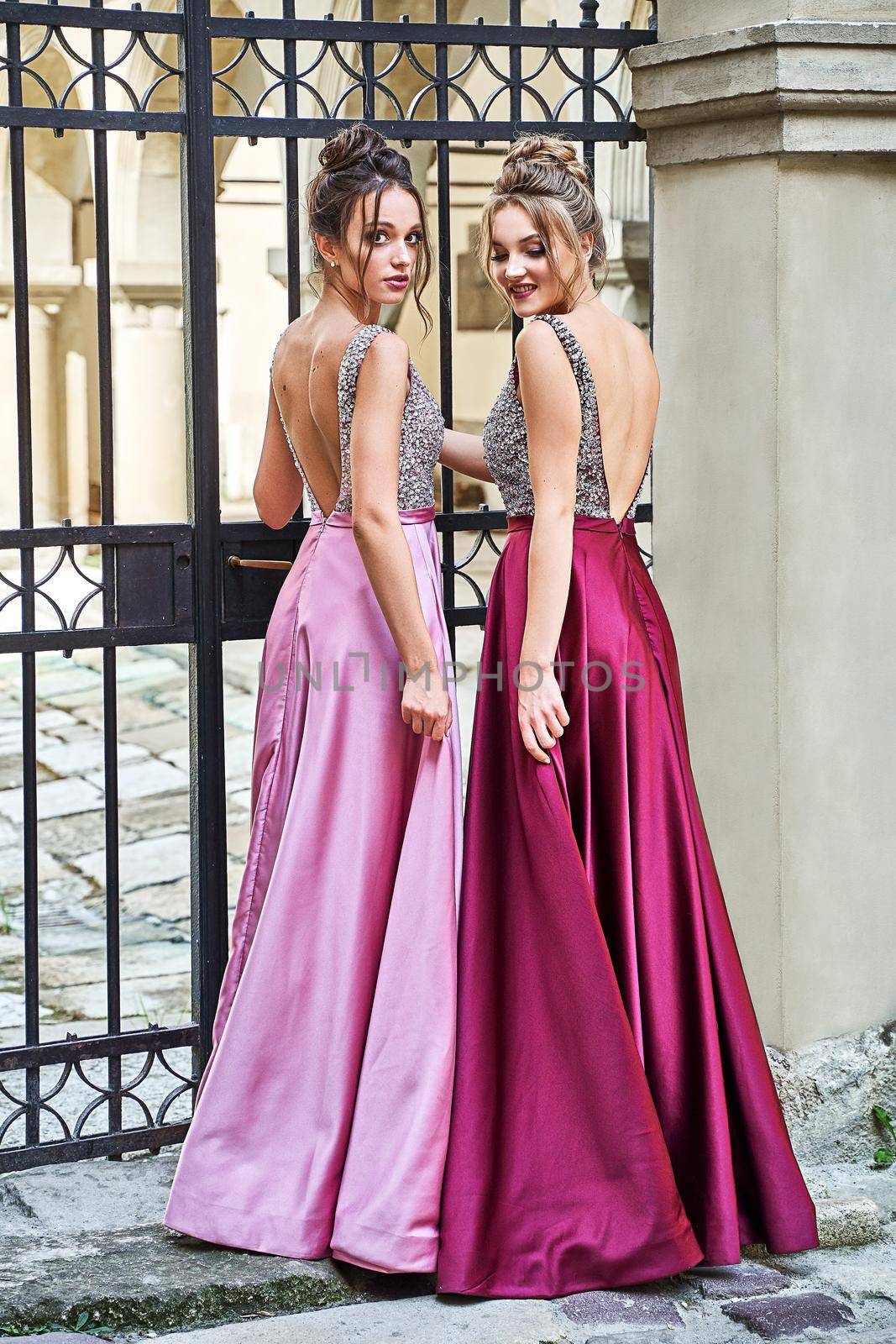 Two beautiful bridesmaids girls blonde and brunette ladies wearing elegant full length purple violet lilac lavender satin bridesmaid dress with silver sequined camisole top. European old town location for wedding day.