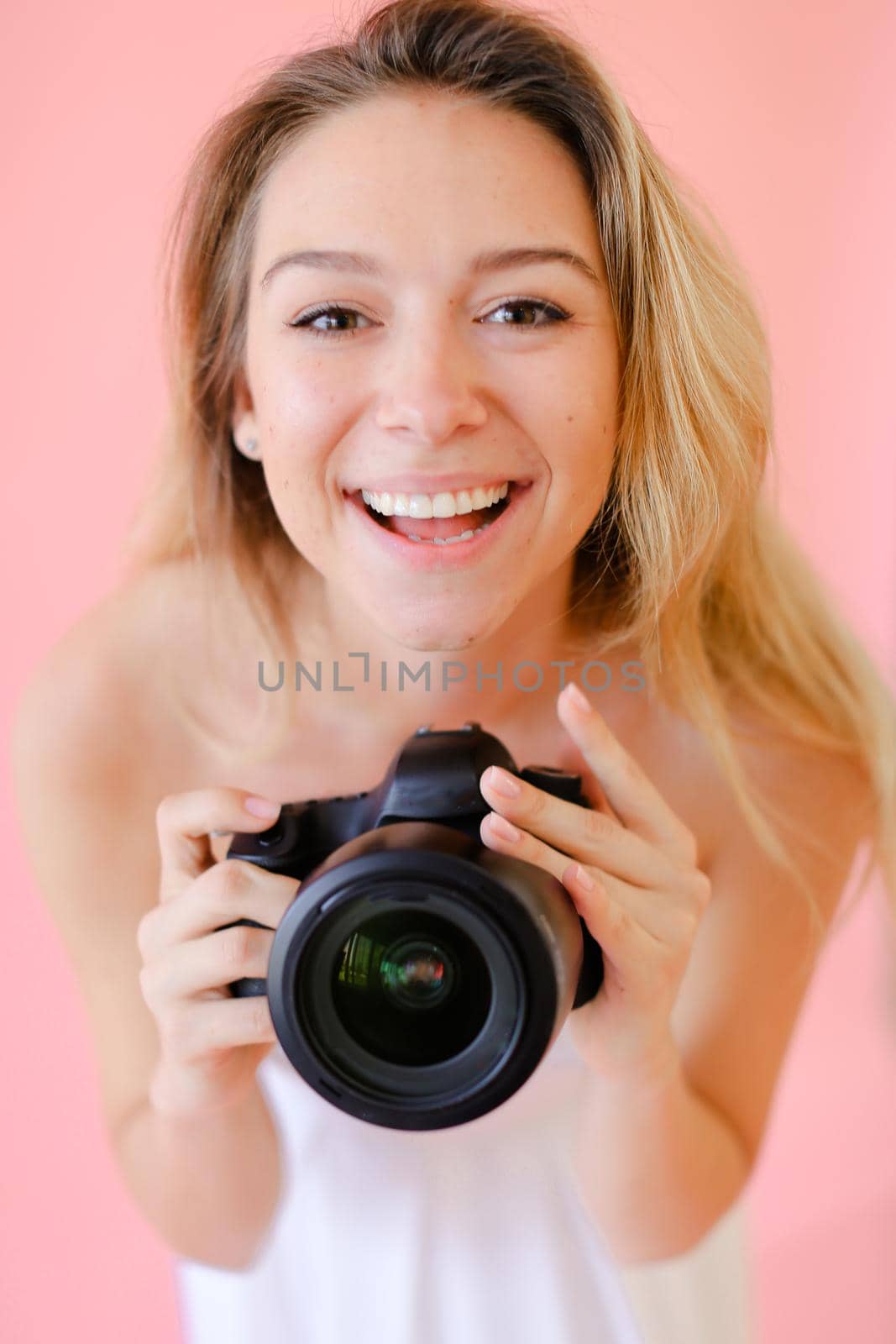 Closeup young caucasian gladden female photographer with black camera in pink monophonic background. Concept of modern profession and photo session.