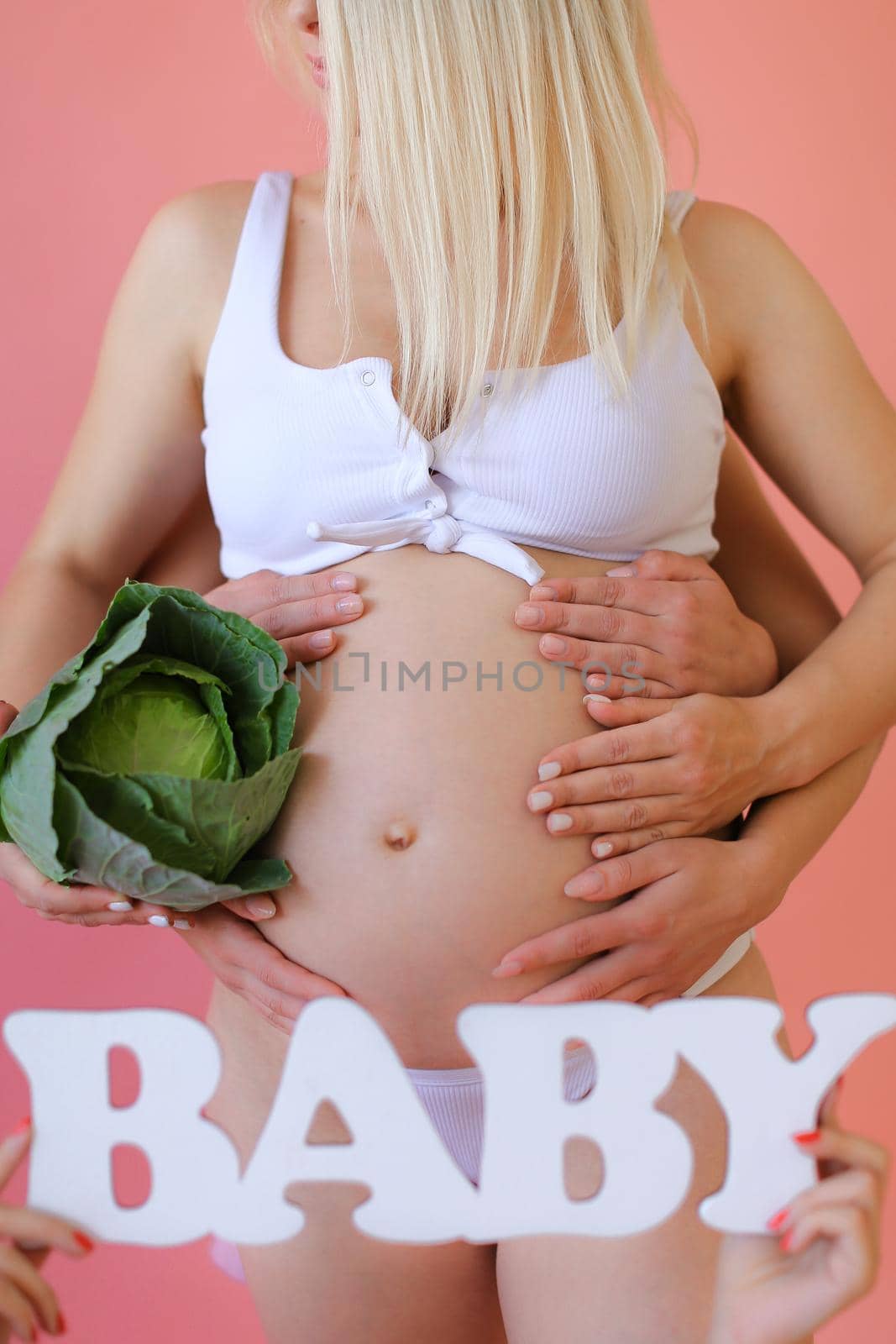Closeup pregnant woman in underwear keeping inscription baby and cabbage in pink monophonic background. Concept of expactant photo session.