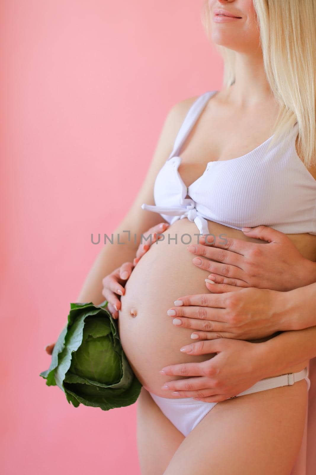 Blonde happy pregnant woman in underwear holding belly and keeping cabbage in pink monophonic background. Concept of expactant photo session.