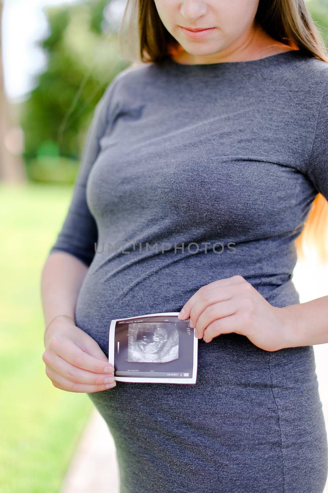 Young happy pregnant girl wearing grey dress keeping ultrasound photo. Concept of expectant female person and motherhood.