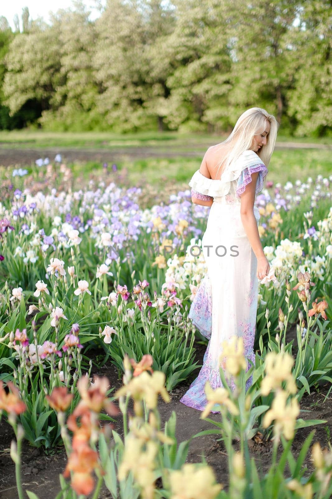 Young blonde woman wearing white dress standing near irises on garden. Concept of human beauty and flora, spring inspiration.
