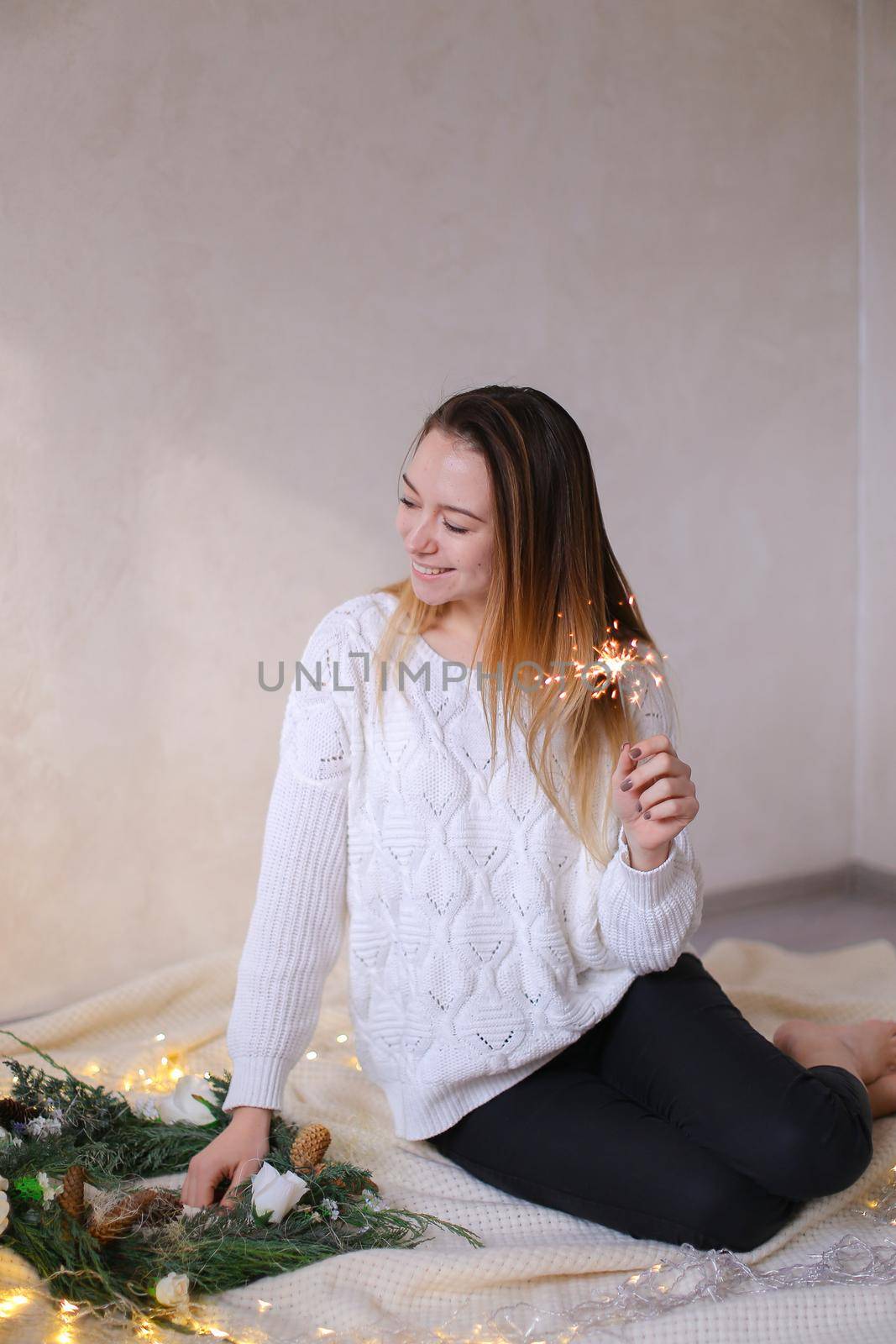 Young caucasian woman sitting with bengal light near twinkling yellow garlands and green wreath. Concept of handmade decorations for Christmas and New Year holidays.