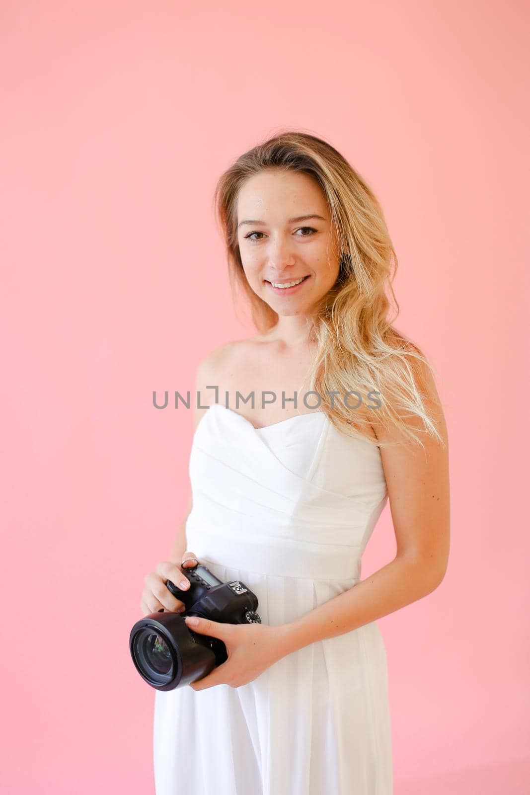 Young european smiling female photographer with camera in pink monophonic background. Concept of modern profession and photo session.