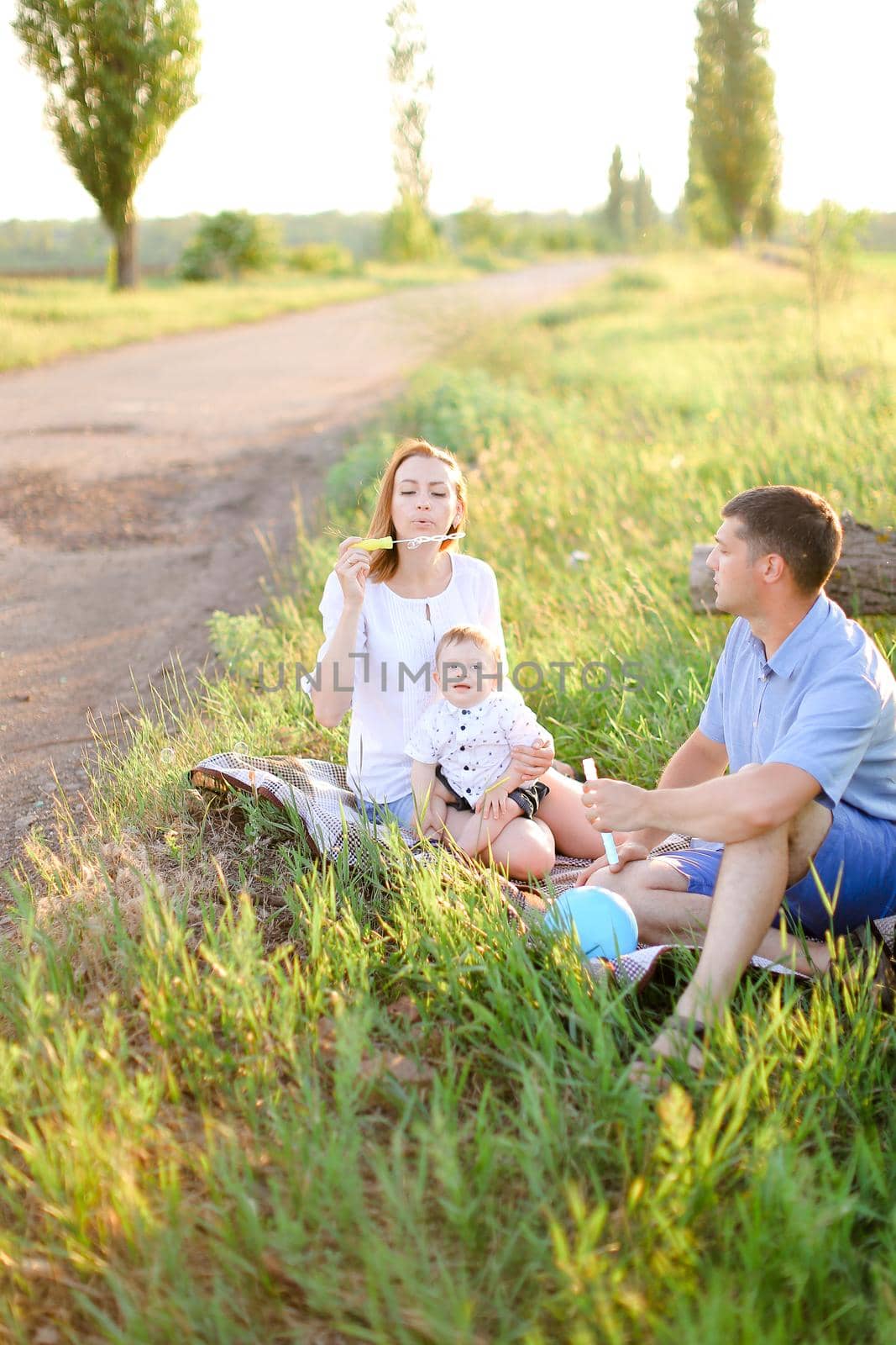 Young happy father and blonde mother sitting on grass with little baby and blowing bubbles. Concept of picnic with child and resting on nature.