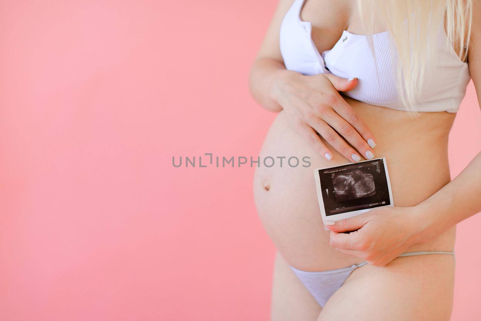 Young happy pregnant woman wearing underwear and holding belly with ultrasound in pink monophonic background. Concept of expectant photo session at studio.