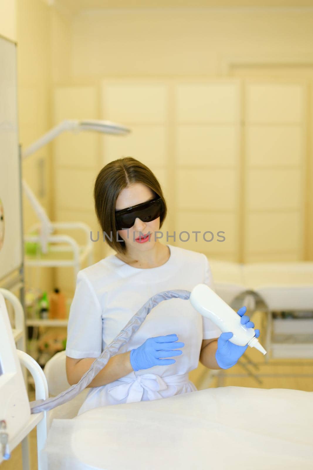 Young skilled cosmetologist wearing special purpose glasses and latex gloves sitting near permanent makeup device. Concept of beauty cosmetology salon and expensive high quality equipment.