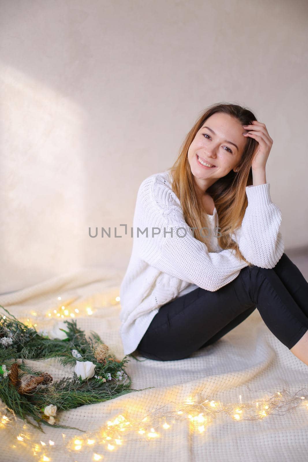 Young happy woman sitting on bed near wreath and yellow garlands. Concept of handmade decorations for Christmas and winter holidays.