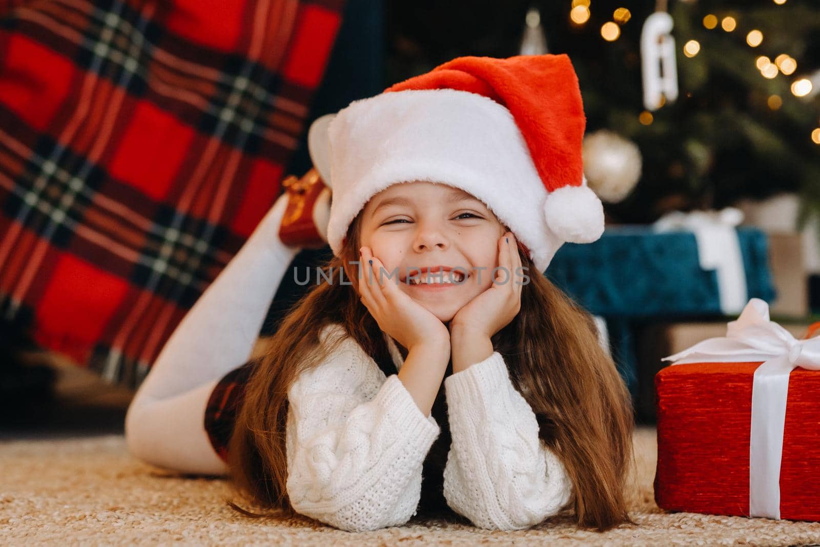 A happy little girl in a Santa Claus hat smiles with gifts in her hands by Lobachad