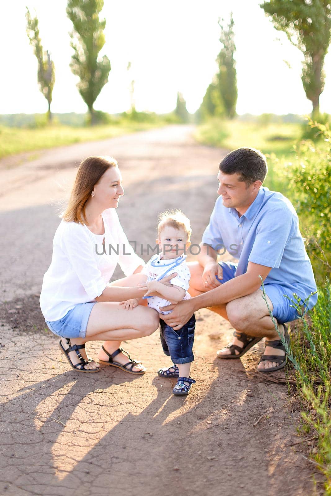 Happy young mother and father sitting on road with little baby, sunshine weather. Concept of parents and children, resting on nature.