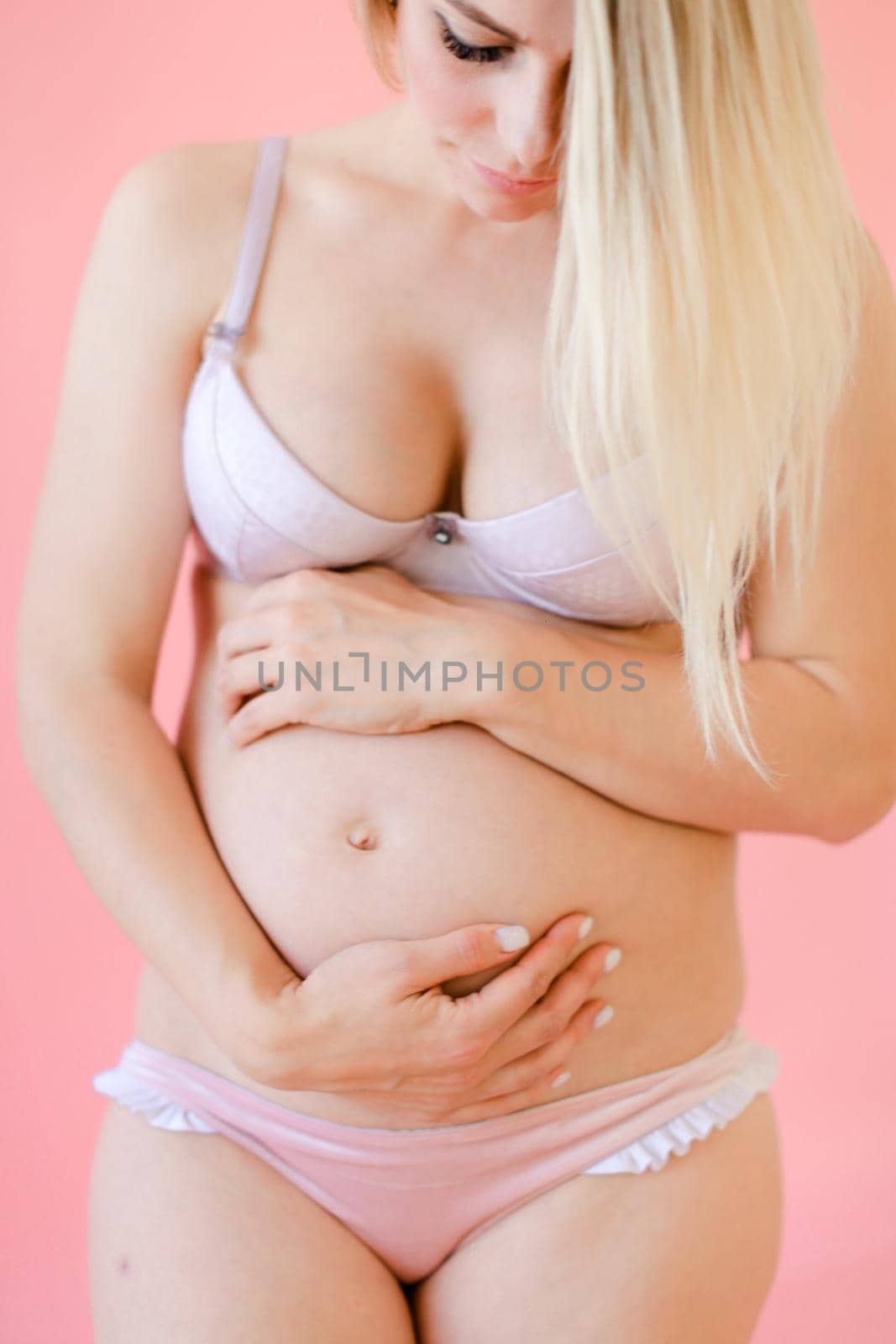 Young pregnant woman wearing underwear and holding belly in pink monophonic background. Concept of expectant photo session at studio.