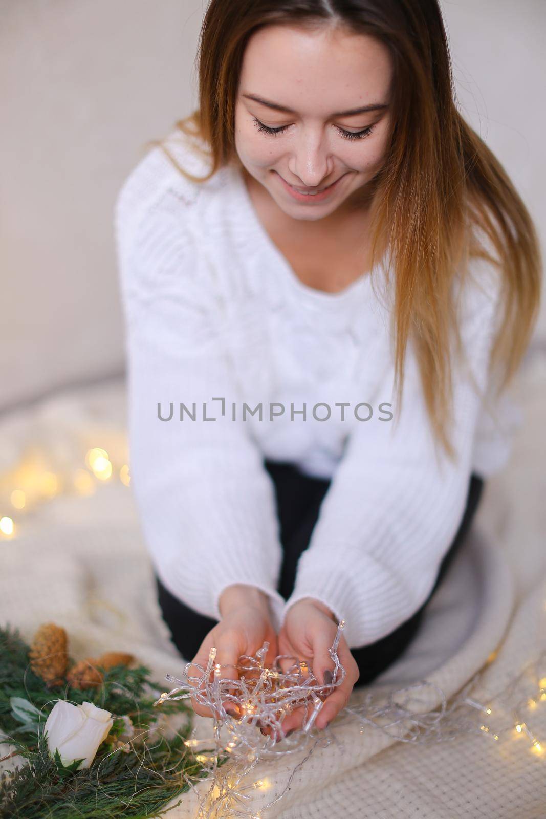 Young woman sitting with twinkling yellow garlands and green wreath. Concept of handmade decorations for Christmas and New Year holidays.