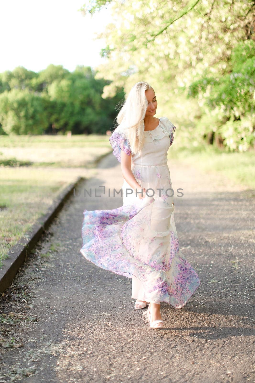 Young blonde woman wearing white dress walking outside near blooming tree. by sisterspro