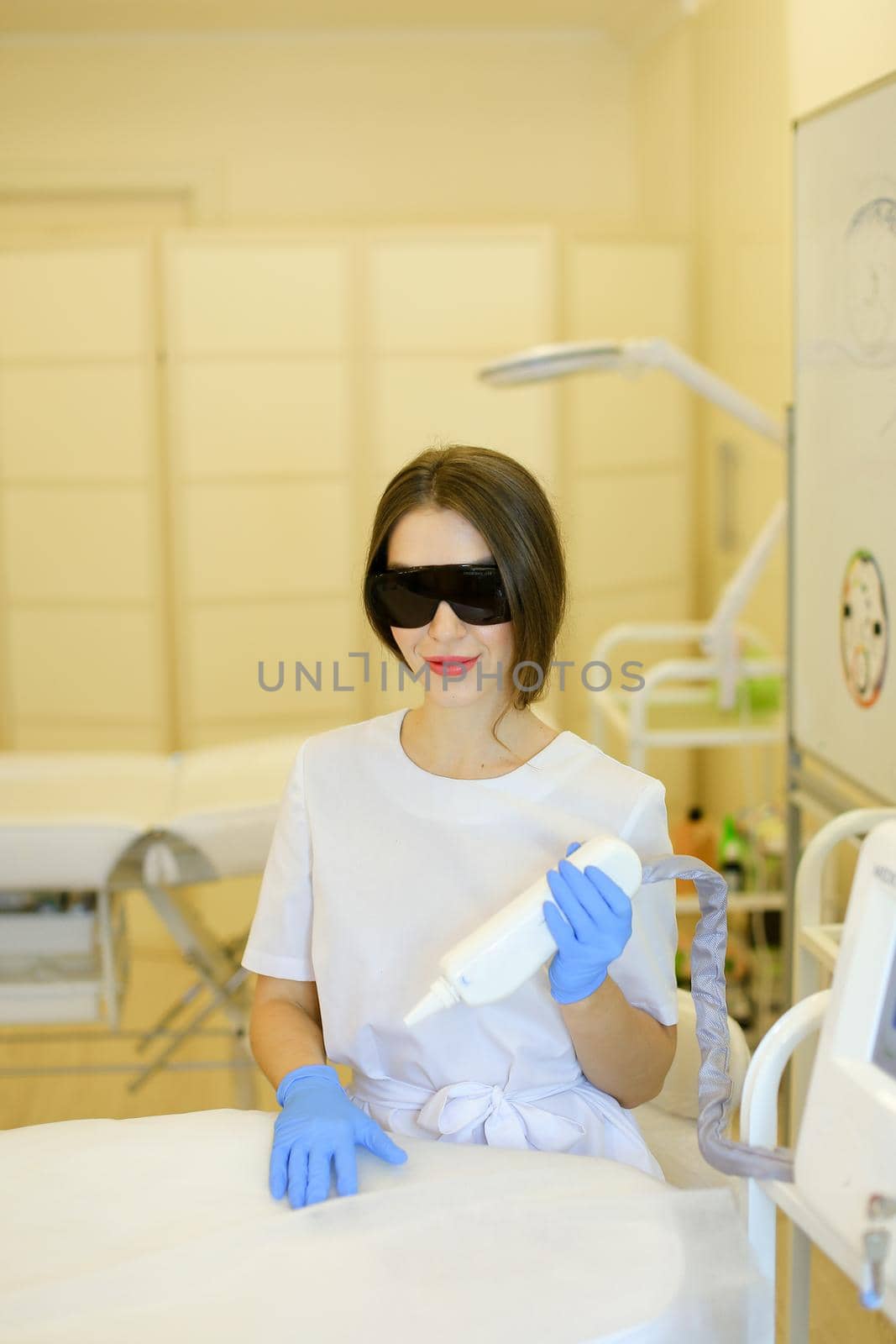 Young caucasian cosmetologist wearing special purpose glasses and latex gloves near permanent makeup device. Concept of beauty cosmetology salon and expensive high quality equipment.