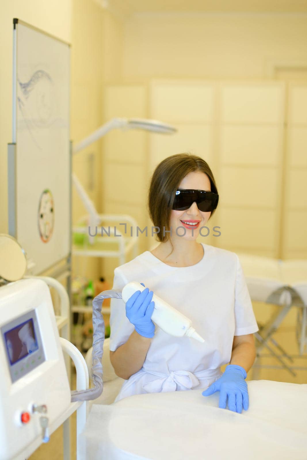 Young caucasian cosmetologist wearing special purpose glasses and latex gloves sitting near permanent makeup device. Concept of beauty cosmetology salon and expensive high quality equipment.