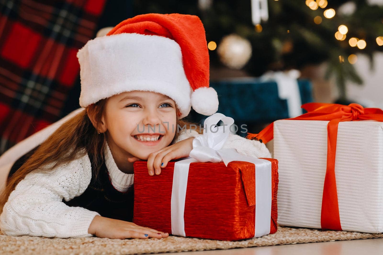 A happy little girl in a Santa Claus hat smiles with gifts in her hands.